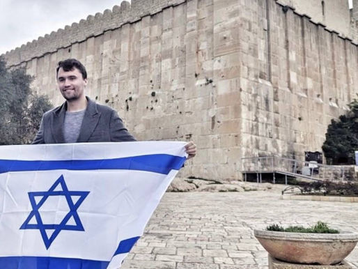 Charlie Kirk stands in front of an ancient stone wall with a minaret visible, holding a large Israeli flag featuring blue stripes and a blue Star of David. The scene is set on a paved courtyard with a potted plant in the foreground, and trees and a ramp are visible in the background.