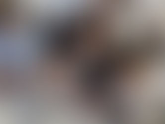 Families and children participate in a hands-on Hanukkah craft activity at a community event. A young woman in a black-and-white patterned dress kneels between two little boys at a white table covered with metal nuts, bolts, glass pieces, glue, and tools. The children, including one wearing a blue kippah and another in a gray sweater, carefully assemble their own miniature menorahs or sculptures. More kids and parents work at nearby tables in a bright, open white-walled space filled with joyful creativity and holiday spirit.