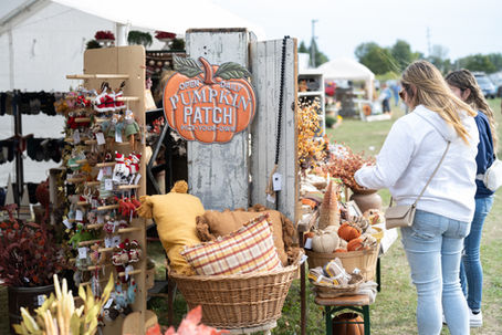 Women browsing autumn items at an outdoor market with a sign reading "Pumpkin Patch." Display features fall decor, cushions, and ornaments.