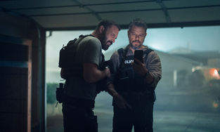 Ben Affleck and Matt Damon wearing police gear and looking at a phone in a garage.