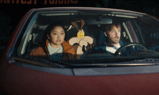 Lana Condor, Wile E. Coyote and Will Forte sitting in a car with shocked or worried expressions on their faces.