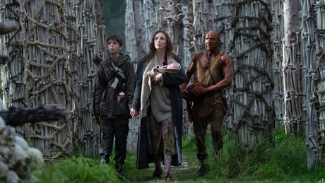 Alfie Williams, Jodie Comer holding a baby, and Ralph Fiennes walking towards and looking up at a memorial composed of human skulls.