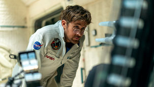 Ryan Gosling, rocking funky glasses and a space uniform, looks at something with curious eyes.