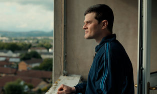 Robert Aramayo standing on a balcony several floors above the ground, looking at and taking in the view.
