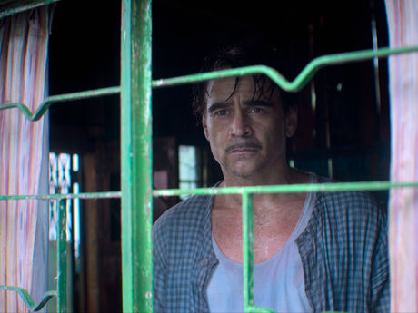 Colin Farrell looking out the window that has green bars on it.