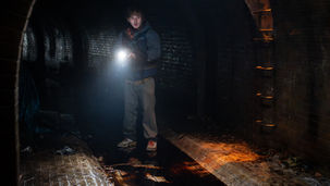 Austin Abrams hanging out in an underground sewer, holding a flashlight.