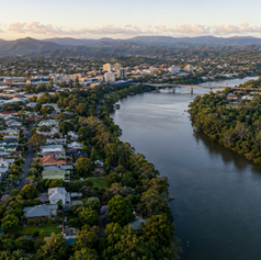 High aerial view of an Australian city beside a broad winding river, with leafy residential neighbourhoods, mature street trees, bridges, dense riverbank vegetation, urban buildings and forested hills in the distance under soft natural light.