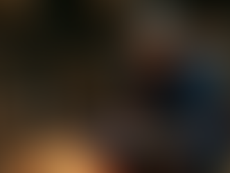 Elderly woman in blue saree writes on a slate in a dimly lit room with stone walls, baskets, and a candle nearby. Calm and focused mood.