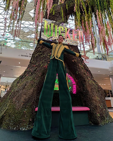 Professional stilt walker wearing a Wicked movie themed green and gold outfit, creating bold visual entertainment and performance artistry for themed events and shows.