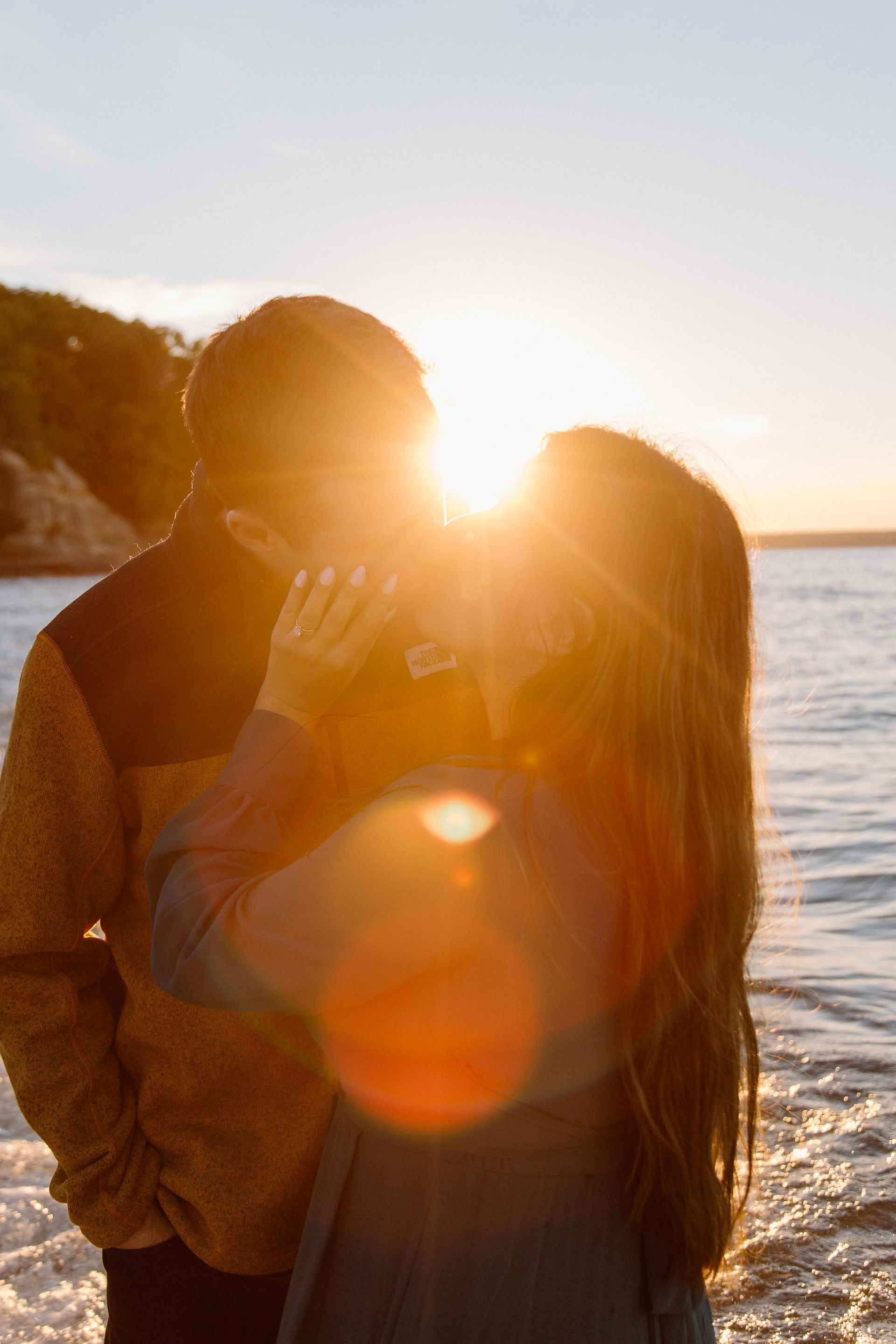 An engaged couple kiss with the sun behind them on the beach in Marquette, Michigan