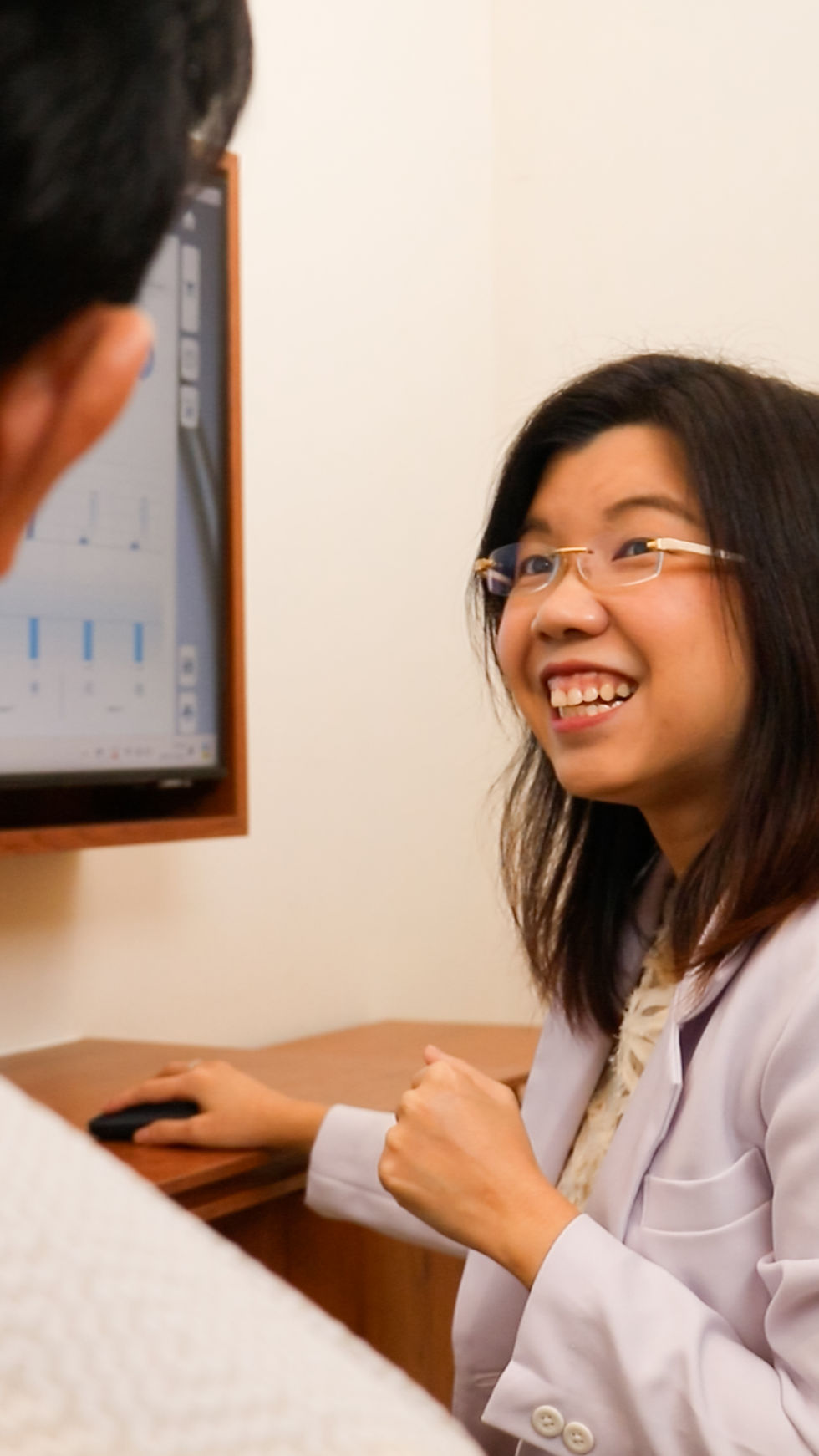 Smiling woman in glasses talks to a person, gesturing towards a screen with charts, in an office setting with a wooden desk.