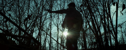 A man standing in the shadows holds a gun in the forest. 