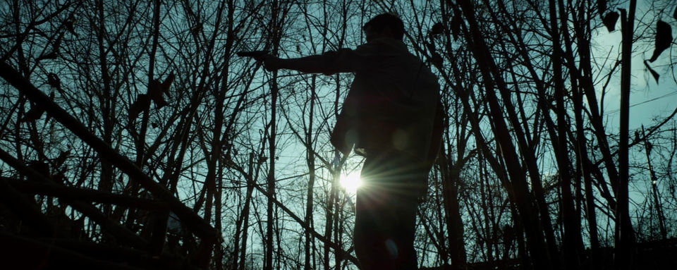 A man standing in the shadows holds a gun in the forest. 