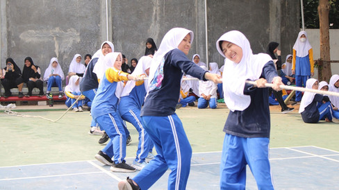 Keseruan dua murid perempuan berhijab putih mengenakan kaos hitam dan celana olahraga biru sedang tertawa sambil menarik tali tambang di barisan depan timnya saat class meeting