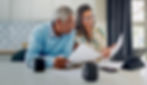 A senior couple sits together in their kitchen while reviewing documents and using a laptop