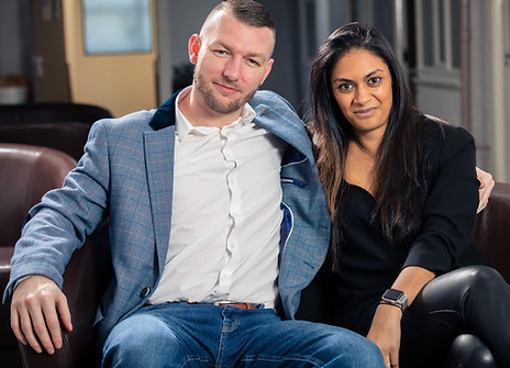 A man in a blue suit jacket & jeans is sat on a sofa with his arm around his partner who is wearing a black shirt and leather pants. They are sat smiling, looking into the camera.