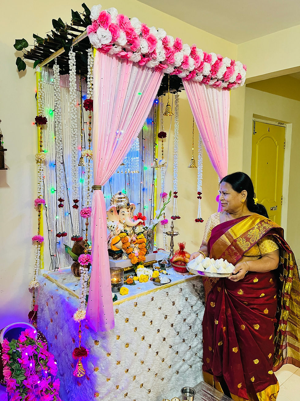 Woman in a maroon sari holds a plate of offerings at a decorated altar with a Ganesha idol, pink drapes, flowers, and colorful lights.