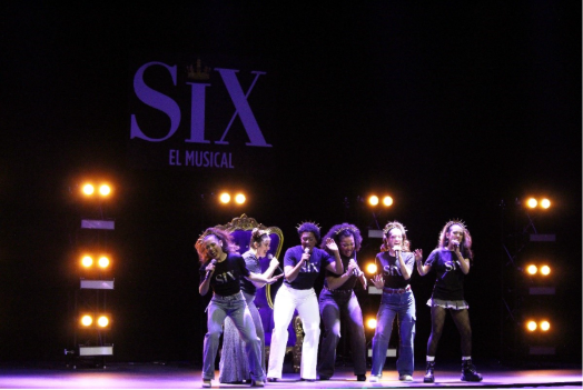Six musical theatre actresses singing into mics on stage