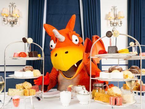A full size Zog the dragon with afternoon tea set out on a table in front of him.