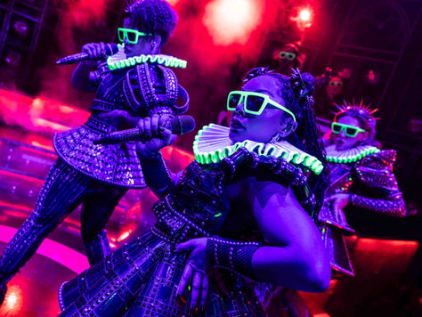 Three women in tutor ruffs and sunglasses all glowing up under UV lights pose on stage
