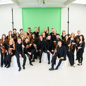 An orchestra of young musicians in black clothes hold their instruments in front of a green paper roll backdrop in a white room