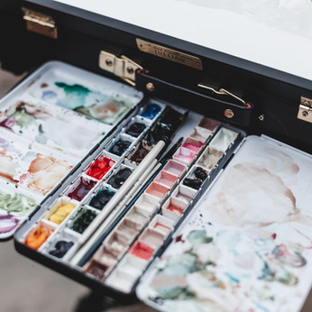 A close up of a paint palette and black easel with an in-progress painting. This artist was painting on sight and creating storytelling art.