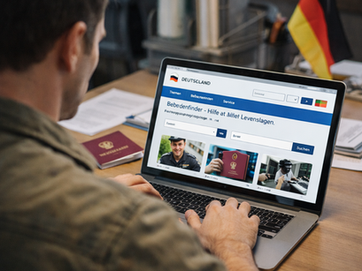 Άντρας κάθεται σε γραφείο και χρησιμοποιεί φορητό υπολογιστή για να βρει πληροφορίες σε γερμανική κυβερνητική ιστοσελίδα, με έγγραφα και διαβατήριο πάνω στο τραπέζι.