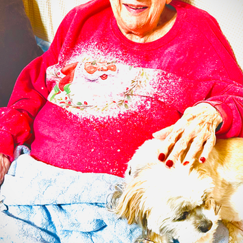 Elderly woman with short gray hair and glasses, smiling while wearing a red Christmas sweater. She is sitting on a couch with a small dog beside her, resting on a blue blanket.