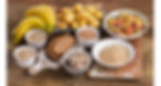 Assorted carbs on a wooden table, including bananas, potatoes, pasta, bread, rice, beans, and grains in bowls. Rustic, colorful display.