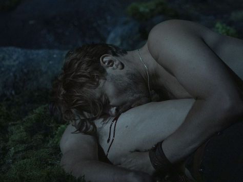 A white man with fair hair is biting on the chest of another white man and drawing blood.