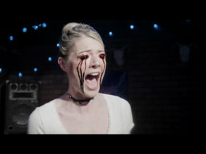 A young white woman with blonde hair tied up in a bun is screaming as her eyes bleed.