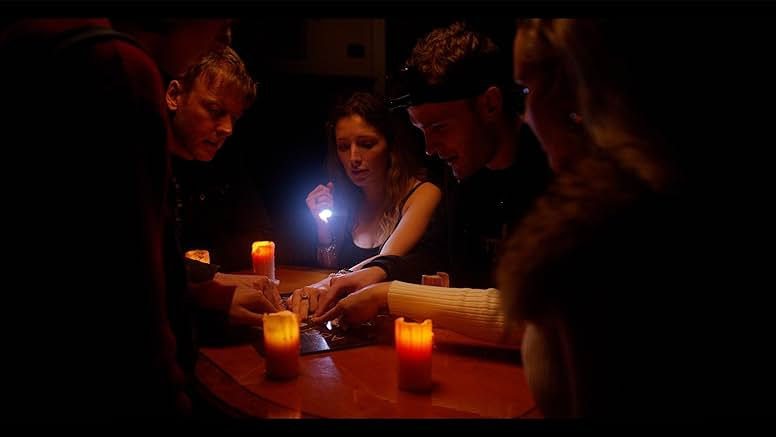 A group of people are sat around a wooden table with lit candles and a ouija board on it. their hands are in the middle on the planchette