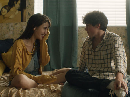 A young teenage white girl is sitting on a bed wearing a yellow shirt. She is sitting opposite a young teenage white boy with dark hair wearing a white check shirt and jeans. They are looking at each other and smiling.