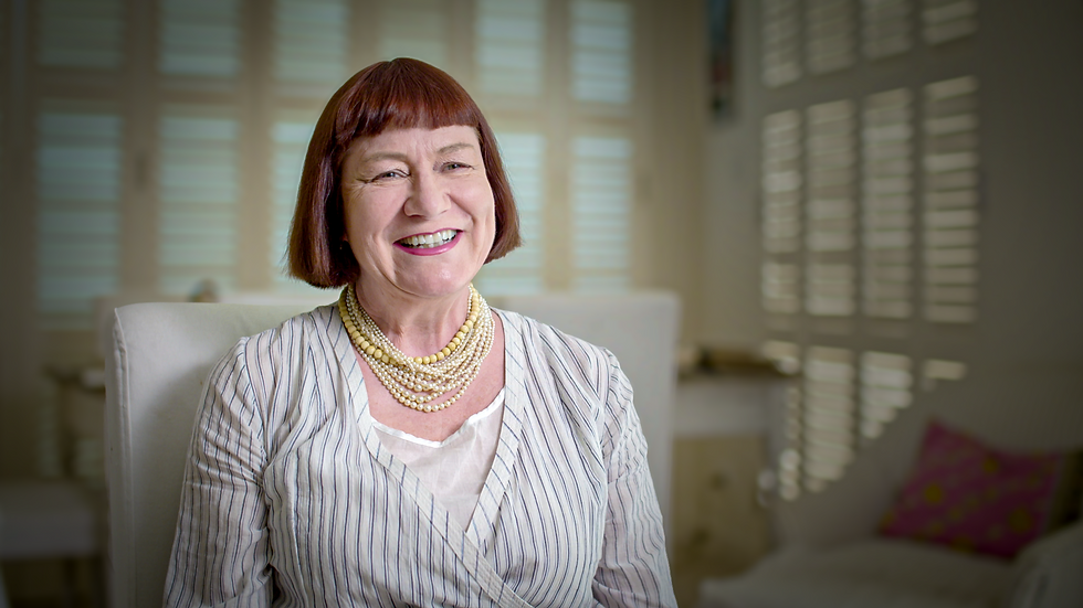 the actress Nell Campbell is a white middle aged woman who has red-brown hair cut into a blunt bob, she is wearing pink lipstick. around her neck are strings of pearls and she is wearing a striped white top