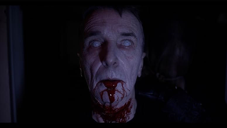 A white middle aged man is staring directly at the camera. His eyes are white and theres blood dripping out of his mouth and down his chin.