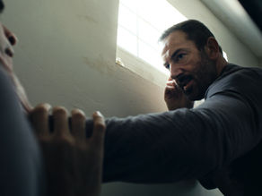 A white man with dark hair and a beard dressed in prison clothes holds a man of Iranian heritage by the throat