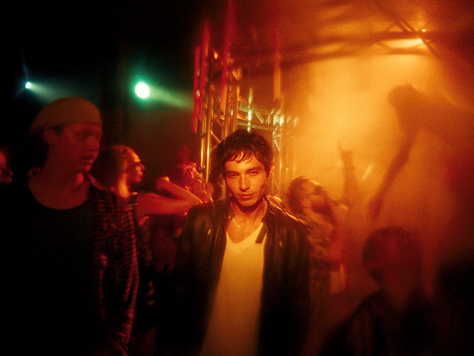 A white man with short dark hair is wearing a black leather jacket and a white tshirt. He is standing in a crowd in the middle of a nightclub.