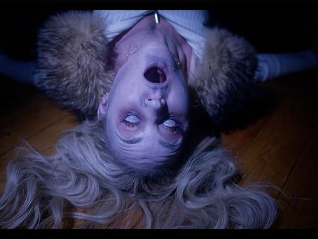 A blonde white woman is lying on a wooden floor with ashy pale skin, her eyes are white and her mouth is wide open