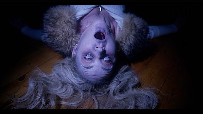 A blonde white woman is lying on a wooden floor with ashy pale skin, her eyes are white and her mouth is wide open