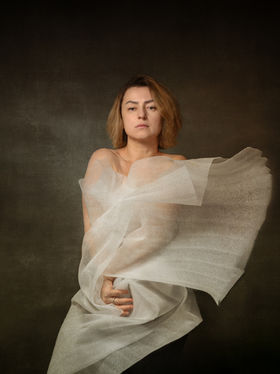 Self-portrait of a woman with translucent fabric gathered around the body, hands subtly engaged, and softened edges, conveying a sense of internal holding and quiet containment.