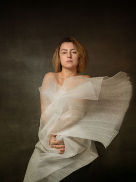 Self-portrait of a woman with translucent fabric gathered around the body, hands subtly engaged, and softened edges, conveying a sense of internal holding and quiet containment.