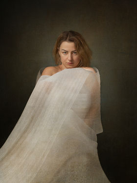 Self-portrait of a woman wrapped in translucent fabric drawn inward around the body, with a direct gaze and a compact posture, suggesting internal concentration and quiet inward movement.