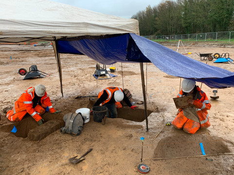 Adapting to the rain. A gazebo and blue tarpaulin cover three archaeologists excavating in the rain.