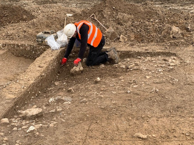 A patch of stones set into orange soil represents the remains of a mettled floor. The floor slopes in the middle indicating something underneath. A man in PPE excavates the stones.