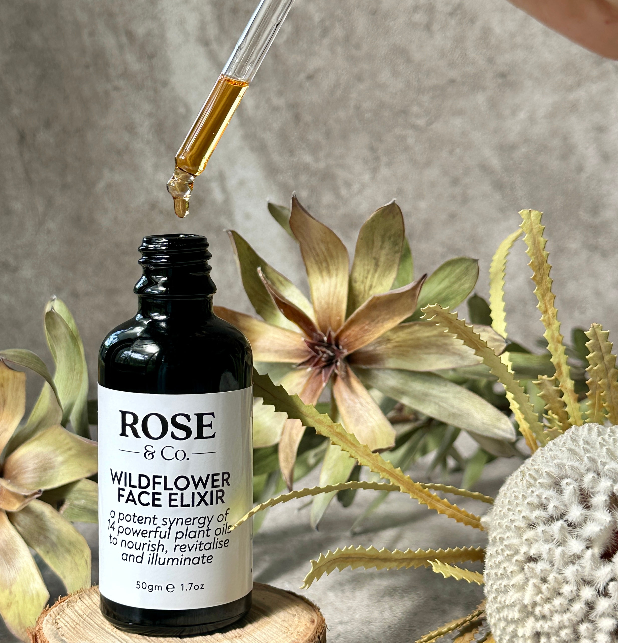 A black glass bottle of face oil with dried native flowers