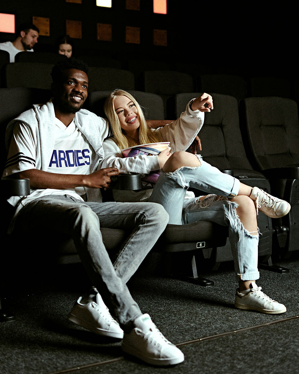 A couple in casual clothes smiles and points while sitting in a dimly lit cinema. The man’s shirt reads "ARDIES." Others are in the background.
