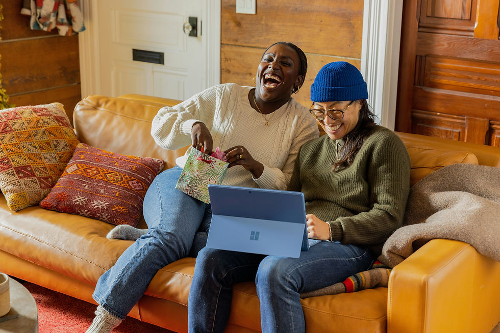Two people laughing on a brown sofa, one holding a gift bag and the other using a laptop. Colorful pillows and a cozy setting enhance warmth.