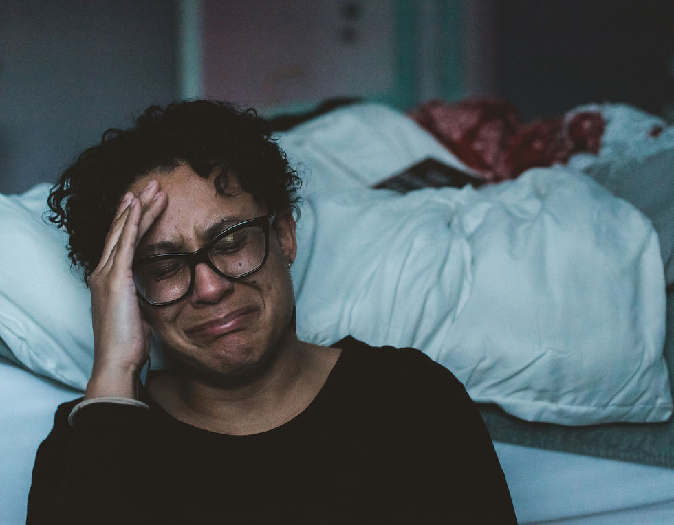 A person wearing glasses is crying, sitting by an unmade bed, hand on head. Dimly lit room with a somber mood, dark clothing.