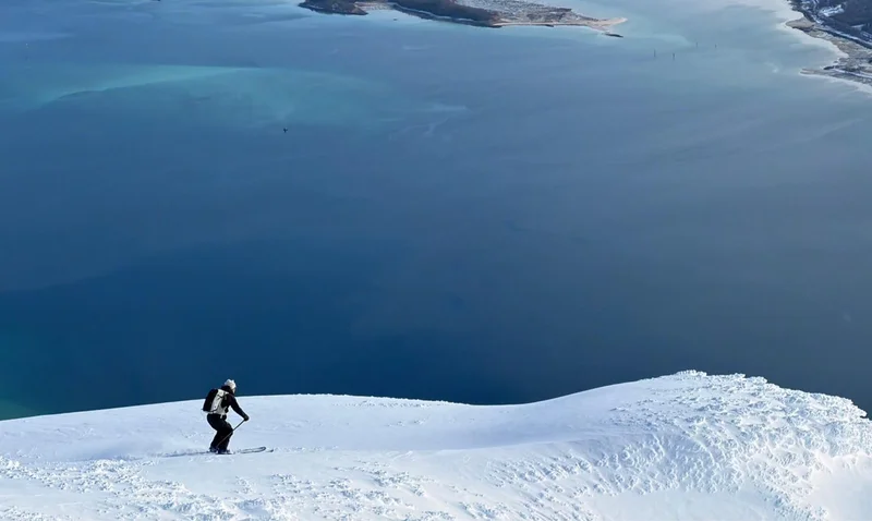 Skieur qui attaque sa descente sur une arête poudreuse au-dessus d'un fjord, Alpes de Lyngen, blog ski PowderWeGo