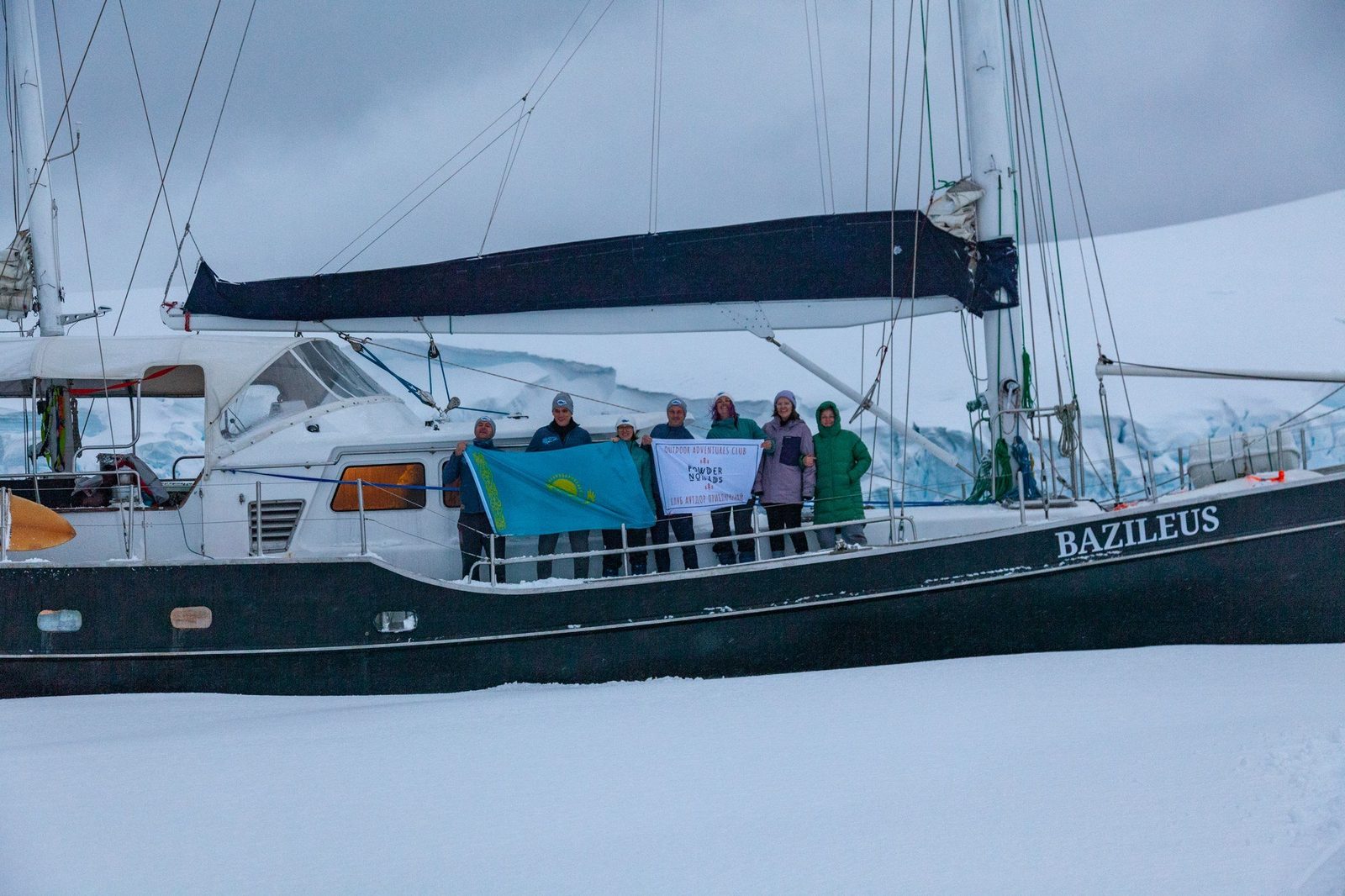 Arktisches Segelschiff vor Anker in einem verschneiten Fjord, schneebedeckte Gipfel dahinter, Basislager der Ski-und-Sail-Reise